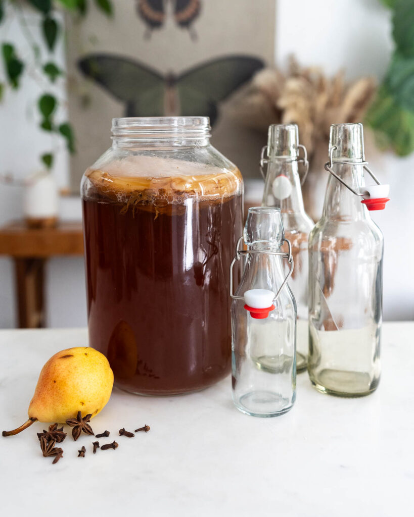 A gallon jar of homemade kombucha sitting beside glass bottles and a pear